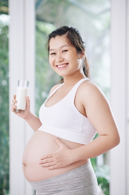 Retrato de feliz joven embarazada sosteniendo un vaso de yogur fresco y tocando su vientre