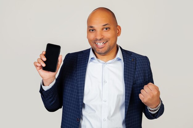 Retrato de feliz joven empresario afroamericano, muestra la pantalla del teléfono inteligente, grita con orgullo y celebra la victoria y el éxito, muy emocionado, se regocija en las emociones.