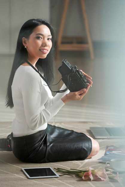 Retrato de feliz joven fotógrafo vietnamita en ropa de blanco y negro sentado en la alfombra y sosteniendo la cámara vintage