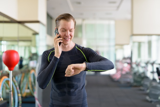 Retrato de feliz joven guapo hablando por teléfono y comprobando smartwatch en el gimnasio durante covid-19