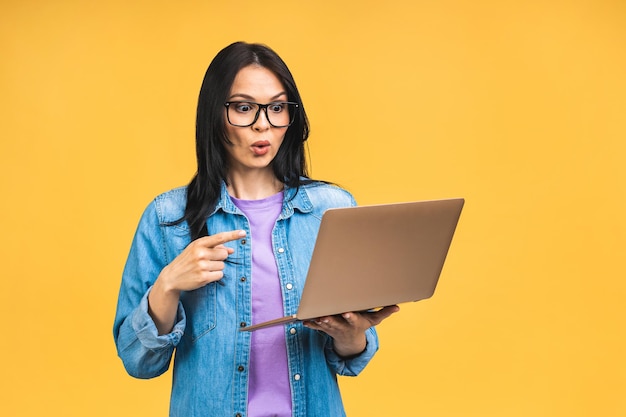 Retrato de feliz joven hermosa mujer sorprendida con gafas de pie con portátil aislado sobre fondo amarillo Espacio para texto