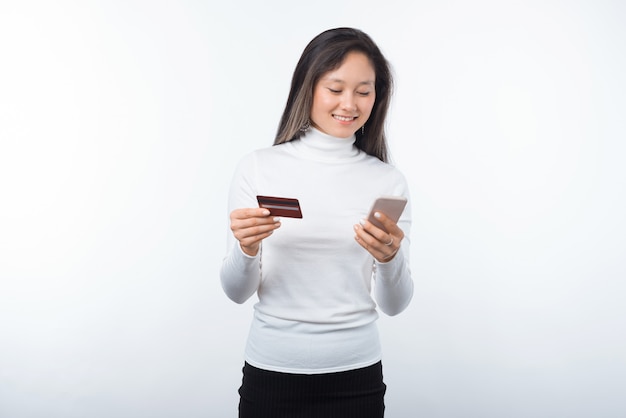 Retrato de feliz joven mujer asiática con tarjeta de crédito y teléfono inteligente sobre fondo blanco.