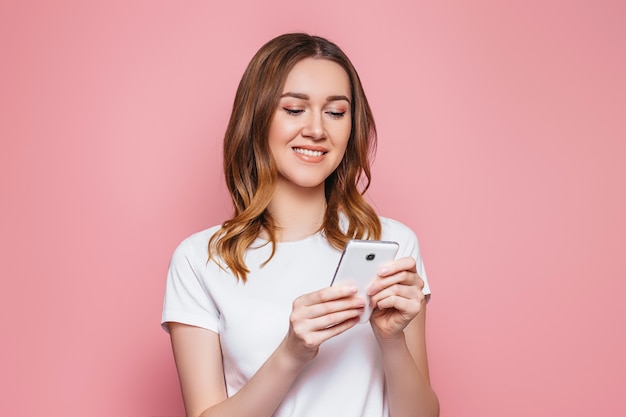 Retrato de una feliz joven mujer cascaica en una camiseta blanca con teléfono móvil aislado sobre pared rosa chica estudiante con cabello ondulado y rizado tiene teléfono inteligente y lee un mensaje