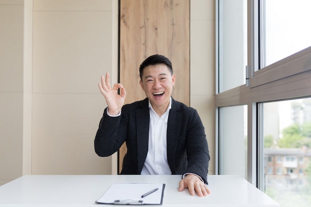 Retrato de feliz joven oficinista masculino asiático mirando a cámara y sonriendo en el interior. Cerca de un gerente amigable con un traje formal y camisa blanca sentado en el trabajo. hombre de negocios sintiendo recibir