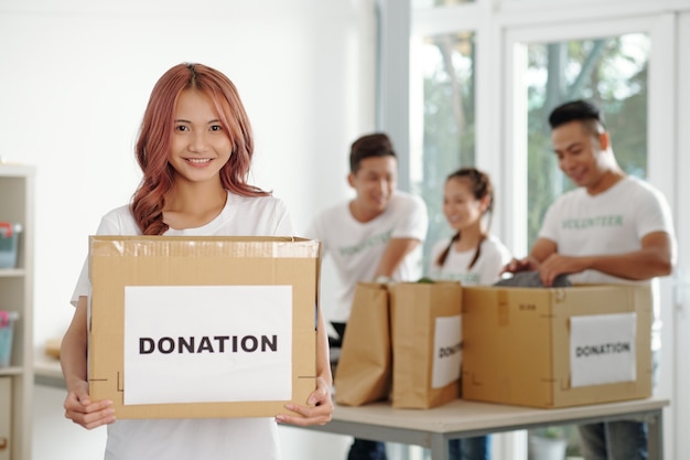 Retrato de feliz joven voluntario de pie en el centro de caridad con cartón de donación en las manos