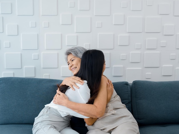 Retrato de feliz madre asiática mayor o abuelo de cabello blanco abrazando a su hermosa hija o nieto sonriendo con amor y comodidad mientras se sienta en un sofá gris en la sala de estar en casa