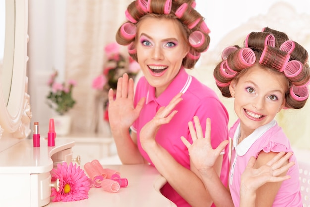 Retrato de feliz madre e hija en rizadores de pelo cerca del espejo