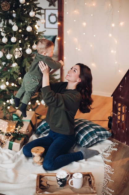 Retrato de feliz madre morena con su bebé en manos divirtiéndose en una manta con bandeja de té caliente y galletas junto al árbol de Navidad decorado festivo