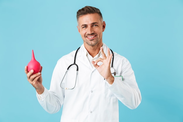 Retrato de feliz médico especialista vistiendo bata blanca sosteniendo enema, que se encuentran aisladas sobre la pared azul