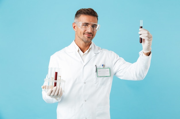 Retrato de feliz médico con gafas sosteniendo tubos de ensayo con sangre, que se encuentran aisladas sobre la pared azul