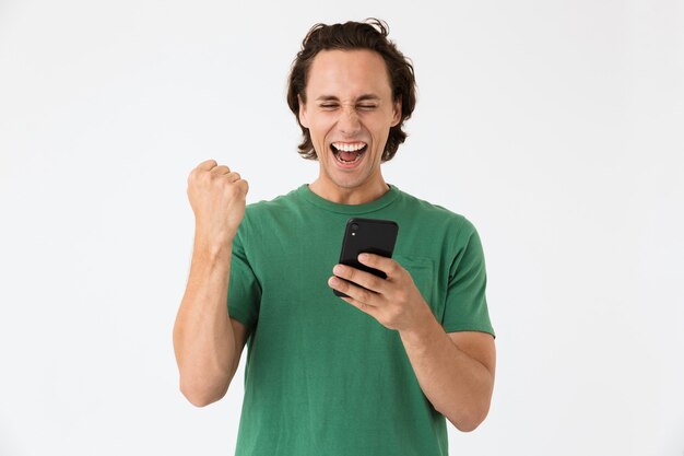 Retrato de feliz morena hombre vestido con camiseta básica gritando y sosteniendo smartphone aislado sobre pared blanca