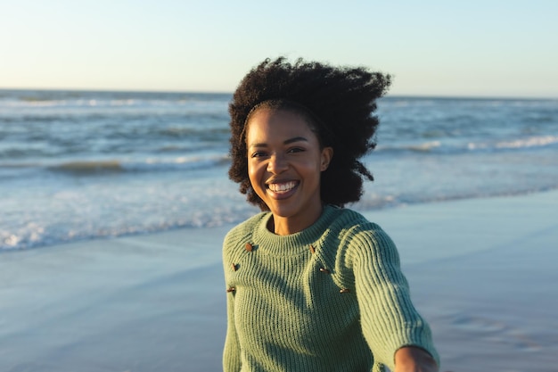 Retrato de una feliz mujer afroamericana mirando la cámara y sonriendo en la playa al atardecer. Verano, relax, tiempo libre y vacaciones, inalterados.