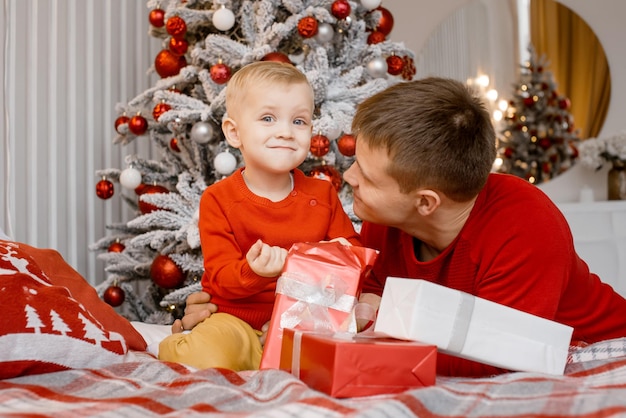 Retrato de un feliz padre e hijo juguetón abriendo un regalo de Navidad cerca del hermoso árbol de Año Nuevo y mirando alegremente a la cámara