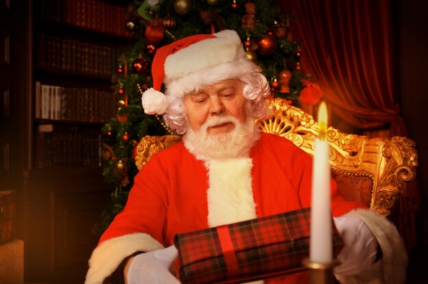 Retrato de feliz Papá Noel sentado en su habitación en casa cerca del árbol de Navidad con caja de regalo.