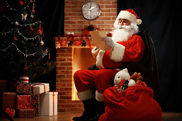 Retrato de feliz Papá Noel sentado en su habitación en casa cerca del árbol de Navidad y un gran saco y leyendo la carta de Navidad o la lista de deseos