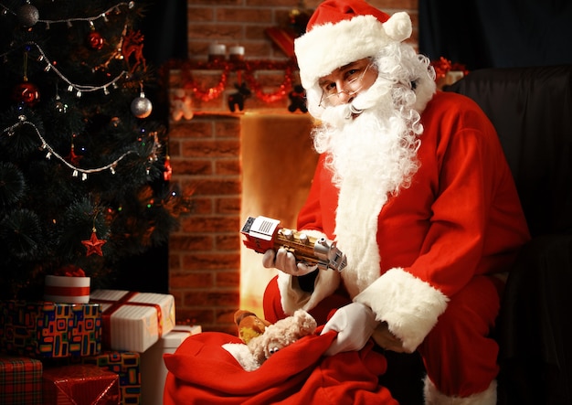 Retrato de feliz Papá Noel sentado en su habitación en casa cerca del árbol de Navidad y un gran saco y leyendo la carta de Navidad o la lista de deseos