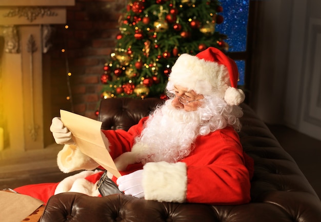 Retrato de feliz Papá Noel sentado en su habitación en casa cerca del árbol de Navidad y leyendo la carta de Navidad o la lista de deseos.