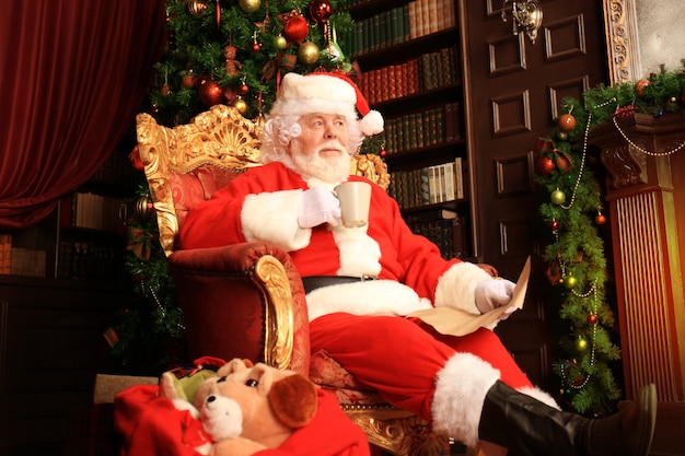 Retrato de feliz Papá Noel sentado en su habitación en casa cerca del árbol de Navidad y leyendo la carta de Navidad o la lista de deseos.