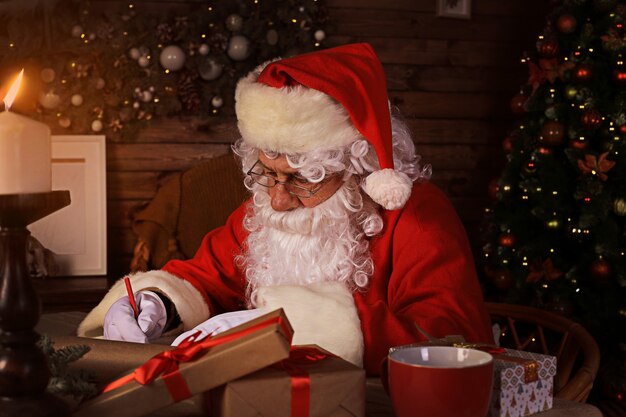 Retrato de feliz Papá Noel sentado en su habitación en casa cerca del árbol de Navidad y respondiendo cartas de Navidad.