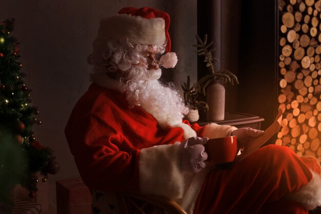 Retrato de feliz Papá Noel sentado en su habitación en casa cerca de la chimenea y leyendo la carta de Navidad o la lista de deseos.