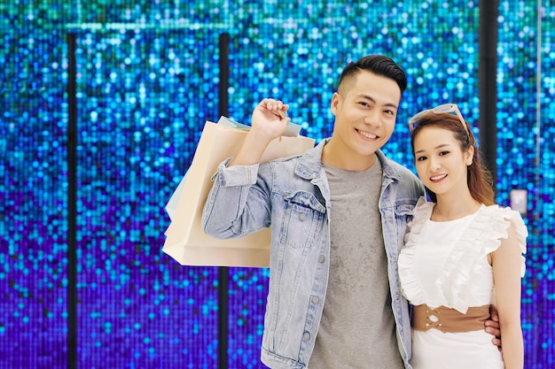 Retrato de la feliz pareja asiática joven de pie en la pared azul brillante con bolsas de la compra y sonriendo al frente
