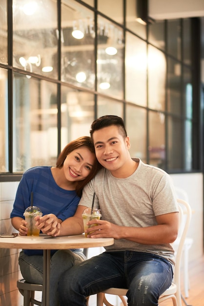 Retrato de la feliz pareja asiática joven sentada con bebidas en una mesa pequeña y tomados de la mano