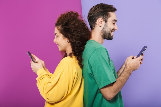 Foto retrato de feliz pareja caucásica en ropa colorida sonriendo y sosteniendo teléfonos celulares de pie espalda con espalda aislado