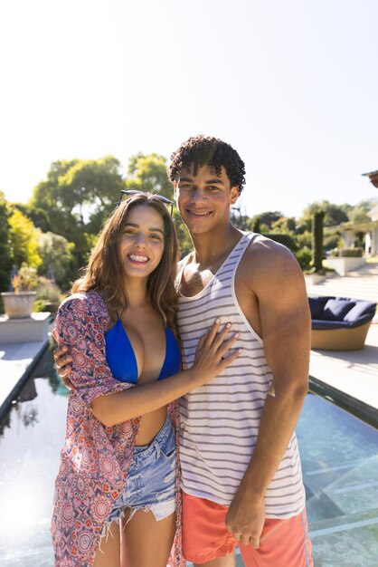 Retrato de una feliz pareja diversa abrazándose y sonriendo junto a la piscina en un jardín soleado, con espacio para copiar. Verano, tiempo libre, relax y vacaciones.