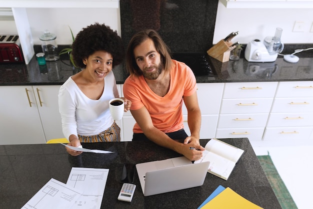 Retrato de una feliz pareja diversa sentada en la cocina usando una computadora portátil y pagando facturas