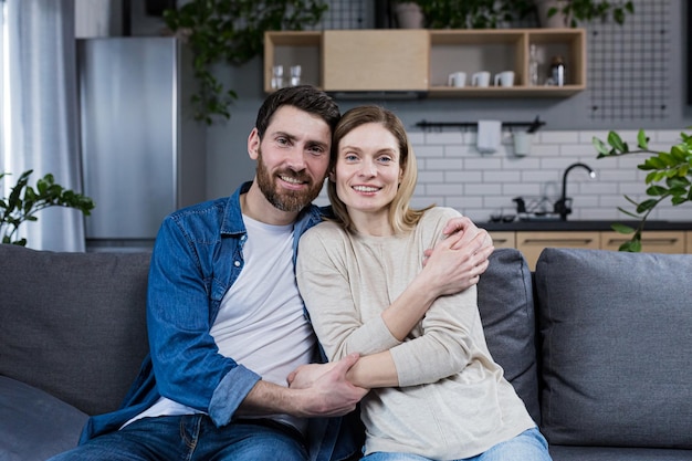 Retrato de una feliz pareja familiar joven Hombre y mujer sentados en el sofá abrazándose saludando sonriendo mirando a la cámara mirándose el uno al otro