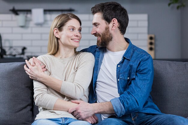 Retrato de una feliz pareja familiar joven Hombre y mujer sentados en el sofá abrazándose saludando sonriendo mirando a la cámara mirándose el uno al otro