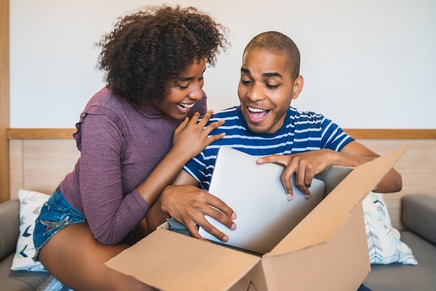 Retrato de la feliz pareja latina joven abriendo un paquete con una computadora portátil dentro, en casa. Concepto de entrega, envío y servicio postal.