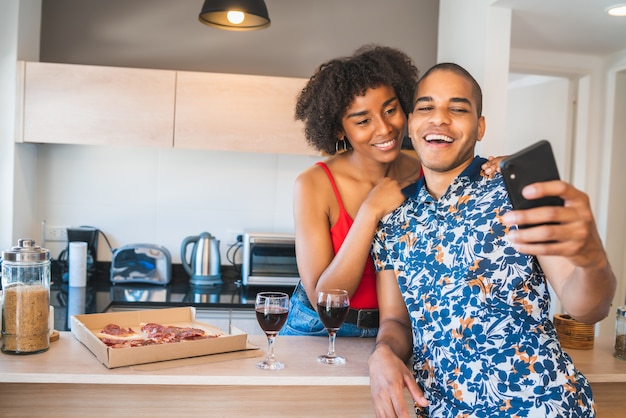 Retrato de feliz pareja latina joven tomando un selfie con teléfono móvil mientras cena en casa nueva. Concepto de estilo de vida y relación.