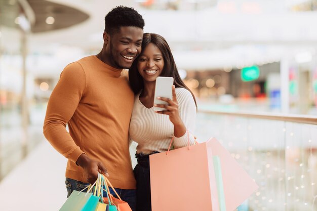 Retrato de feliz pareja negra usando teléfono con bolsas de compras