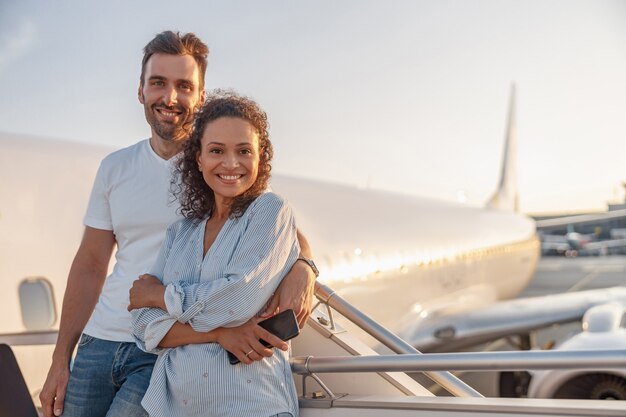 Retrato de feliz pareja de turistas, hombre y mujer mirando emocionados mientras están parados juntos al aire libre listos para abordar el avión al atardecer. Vacaciones, estilo de vida, concepto de viaje