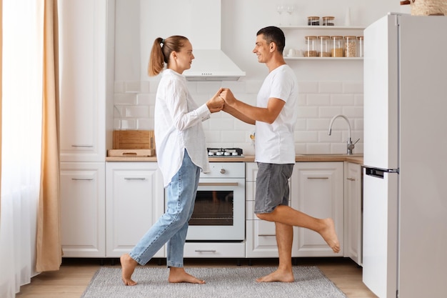 Retrato de feliz positivo joven adulto de raza caucásica hombre y mujer, posando en interiores, bailando en la cocina, celebrando su aniversario, vistiendo atuendos de estilo casual.