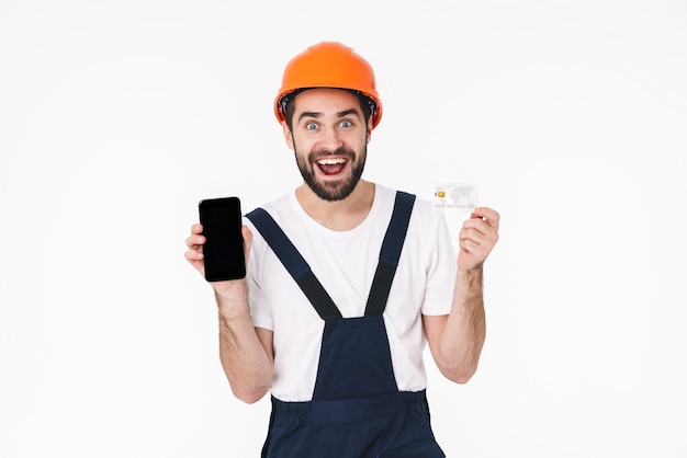 Retrato de feliz positivo joven constructor en casco aislado sobre pared blanca con tarjeta de crédito que muestra la pantalla del teléfono móvil.