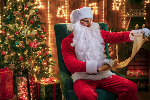 Retrato de feliz Santa Claus sentado en su habitación en casa cerca del árbol de Navidad y leyendo la carta de Navidad o la lista de deseos