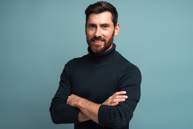 Retrato de feliz satisfecho apuesto joven de pie con los brazos cruzados y mirando a la cámara con una gran sonrisa. Foto de estudio en interiores sobre fondo azul espacio de copia