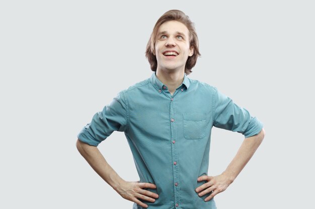 Retrato de feliz soñador guapo rubio joven de pelo largo en camisa casual azul de pie con las manos en la cintura y mirando a otro lado y soñando. tiro del estudio de interior, aislado en fondo gris claro.