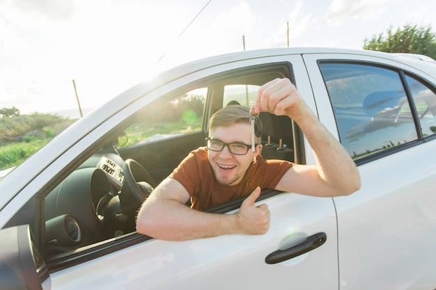 Retrato de feliz sonriente joven, comprador sentado en su coche nuevo y mostrando las llaves fuera de la oficina del distribuidor.