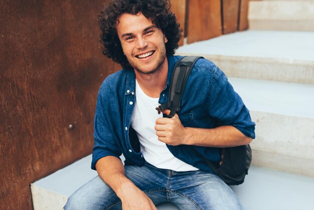 Retrato de feliz sonriente joven estudiante con mochila sentado en las escaleras de hormigón en la calle de la ciudad