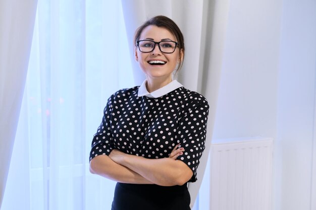 Retrato de una feliz y sonriente mujer de negocios madura con gafas con los brazos cruzados, una mujer mirando la cámara, dentro de una ventana con cortinas