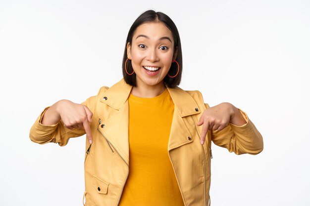 Retrato de una feliz y sonriente niña asiática señalando con el dedo hacia abajo y mostrando el logotipo que muestra una pancarta de pie con una chaqueta amarilla contra el fondo blanco Copiar espacio