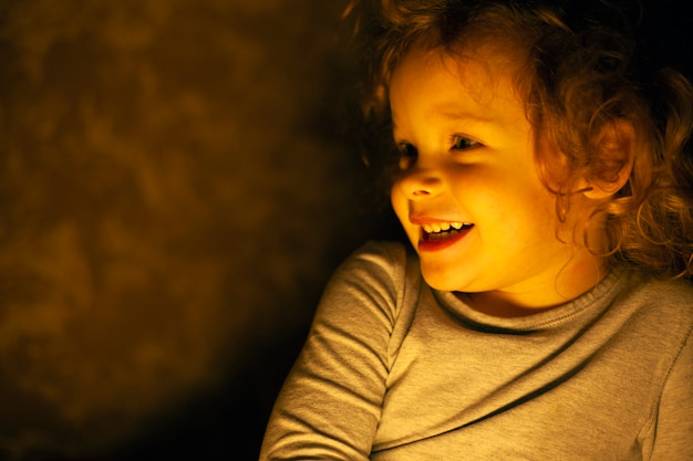 Retrato de una feliz y sonriente niña pelirroja en los cálidos rayos amarillos de una lámpara en el cuarto oscuro.