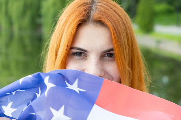 Retrato de feliz sonriente niña pelirroja escondiendo su rostro detrás de la bandera nacional de Estados Unidos