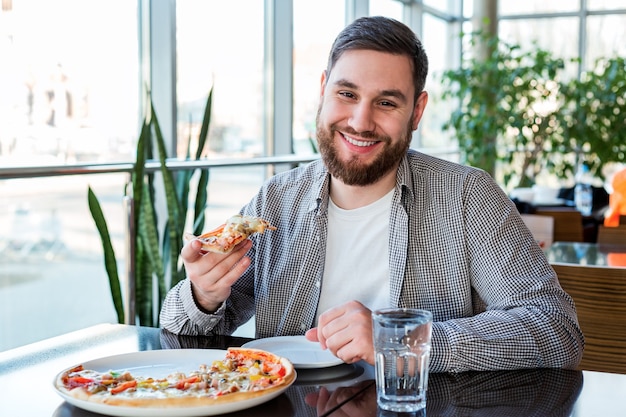 Retrato feliz sorridente homem caucasiano comendo pizza italiana na pizzaria mantém distância social. pizza deliciosa no café.