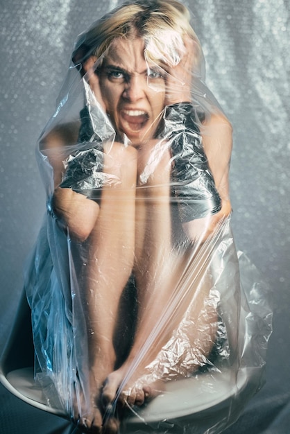 Retrato femenino atrapado. Ataque de pánico. Abuso de la violencia. Mujer gritando desenfocada abrazando la cabeza cubierta por una película de plástico transparente aislada en un desenfoque gris.