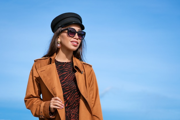 Retrato femenino de una joven y bella mujer con una gorra negra contra un cielo azul con gafas de sol y una chaqueta