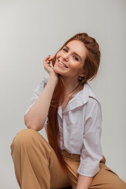 Retrato femenino natural de una hermosa mujer linda alegre con una hermosa sonrisa en ropa de moda con pantalones y una camisa se sienta sobre un fondo blanco en el estudio